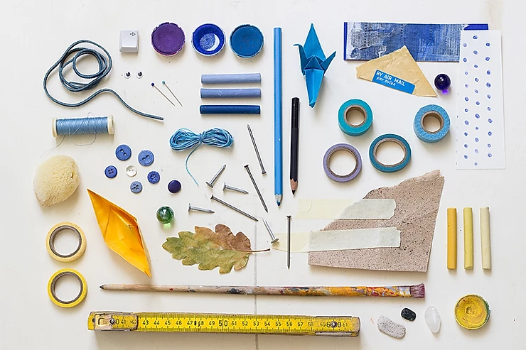 Assorted art and craft supplies, stationery, buttons, tape, thread, nails, leaves, a ruler, folded paper boats, and paintbrush arranged neatly on a white surface.