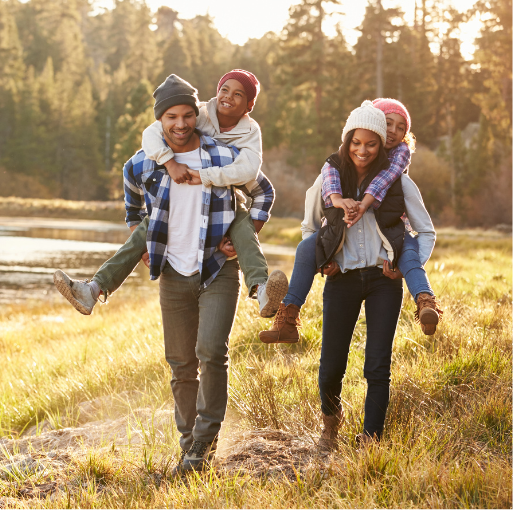 A smiling man and woman give piggyback rides to two kids while walking through a grassy field near a lake and trees, all wearing casual clothes and beanies on a sunny day.
