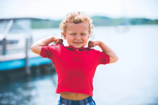 A young child in a red shirt smiles and flexes their arms, showing off their muscles, while standing outdoors near water on a sunny day.