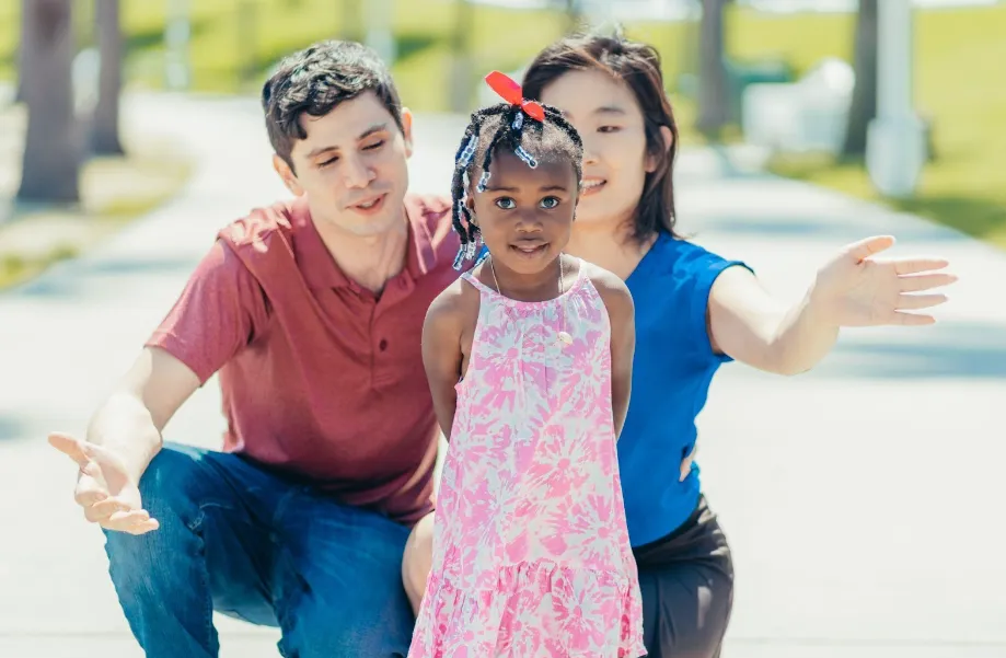 A young girl in a pink dress stands in front of two adults on a sunny day. The adults, one in red and one in blue, kneel behind her with open arms, smiling and encouraging her.