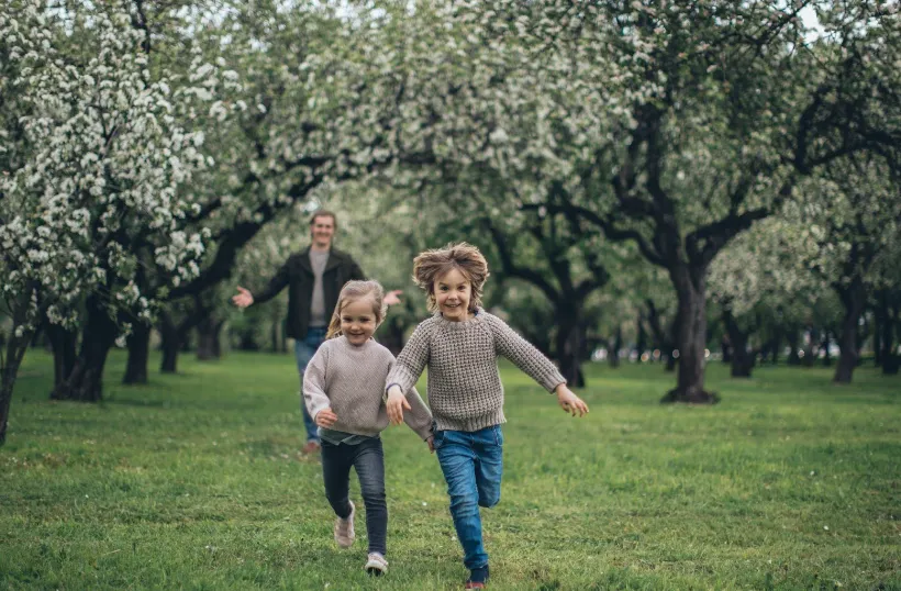 Two children run happily through a green orchard with blooming trees, while an adult follows behind them, smiling. The scene is lively and full of springtime energy.