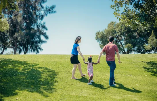 A family of three—two adults and a child—walks hand-in-hand up a grassy hill in a sunny park, surrounded by trees and blue sky.