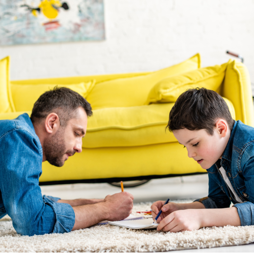 An adult and a child lie on a rug, drawing together on paper. A bright yellow couch is in the background. Both appear focused and engaged in the activity.