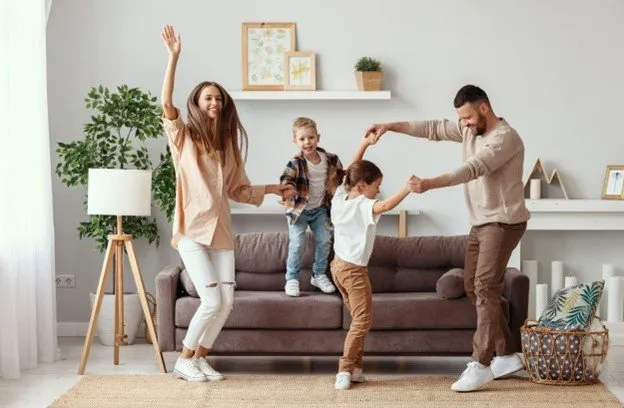 A family of four dances together in a living room. Two adults hold hands with a child while another child stands on the couch, all smiling and appearing joyful. Plants and framed art decorate the bright, cozy space.