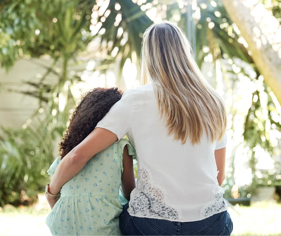 A woman with long blonde hair hugs a young girl with curly hair as they sit together outdoors, facing away, surrounded by lush green plants and sunlight.