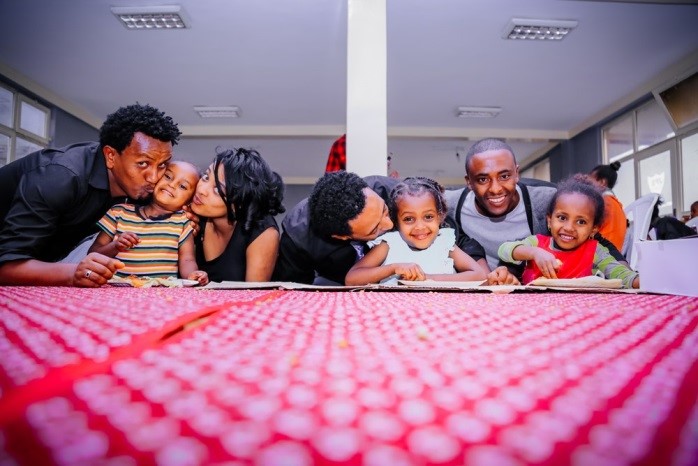 Six adults and children smile and lean over a red tablecloth in a brightly lit room, showing affection and playful expressions. The scene captures a joyful, family-like gathering.
