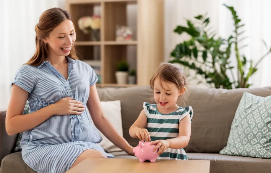 A pregnant woman smiles while sitting on a couch, watching a young girl put coins into a pink piggy bank on a table in front of them. The room is bright with plants and decorative pillows.