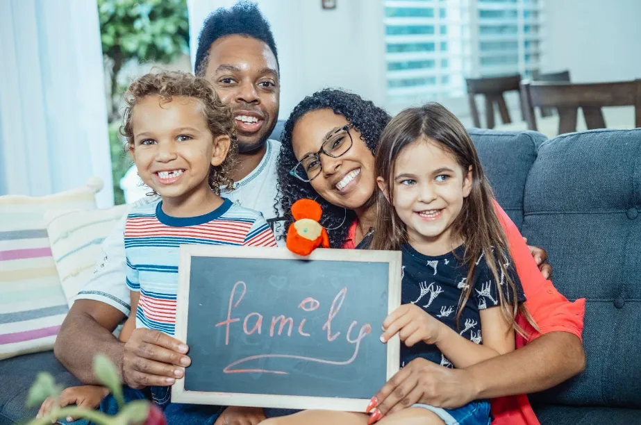 A smiling family of four sits together on a couch, holding a chalkboard with family written on it in cursive. Two children sit in front, and two adults are behind them. They appear happy and close.