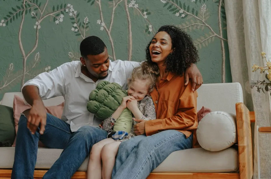 A smiling family sits on a couch; an adult couple hugs a laughing child holding a green pillow. The background features green floral wallpaper and part of a beige curtain.