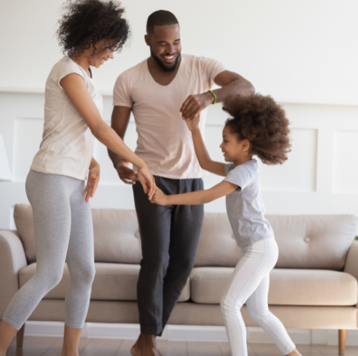 A smiling family of three, two adults and a child, dance together in a living room with a couch in the background. They appear joyful and are holding hands.