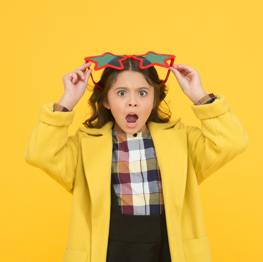 A surprised child in a yellow coat and plaid shirt holds up large, red star-shaped sunglasses against a bright yellow background.