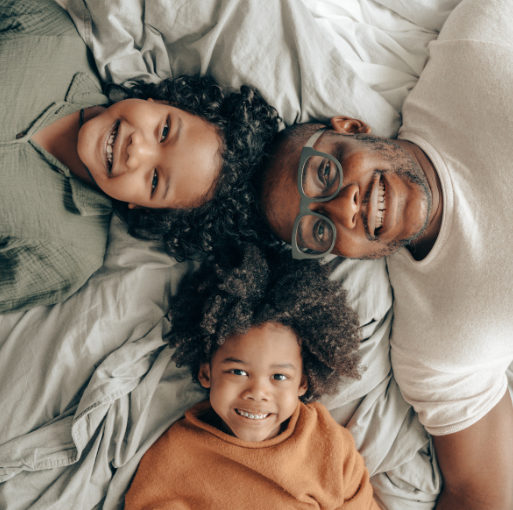 A man and two children lying on a bed, smiling up at the camera. They are close together on a light-colored blanket, creating a warm and joyful atmosphere.