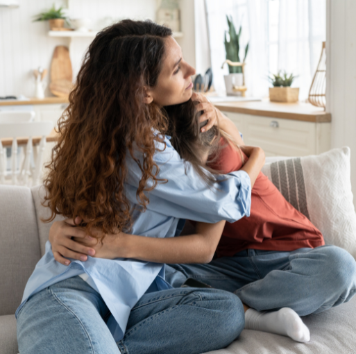Two people sit on a couch in a cozy, sunlit living room, hugging each other closely. One person wears a blue shirt and jeans, while the other wears a rust-colored top and jeans.