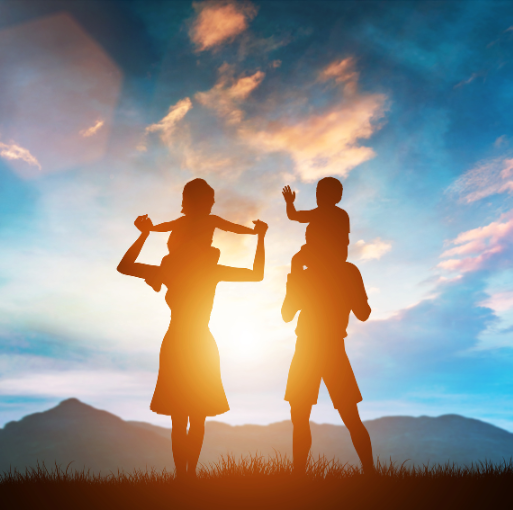 Silhouettes of two adults carrying children on their shoulders stand on grass at sunset, with a bright sky and mountains in the background.