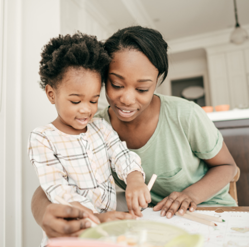 A woman and a child sit together at a table, smiling and drawing on paper with colored pencils in a bright kitchen.