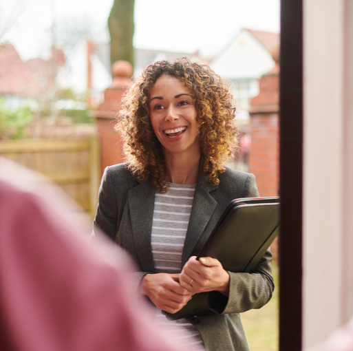 A woman with curly hair, wearing a blazer and striped shirt, smiles while holding a black portfolio at an open door, with houses and a wooden fence in the background.