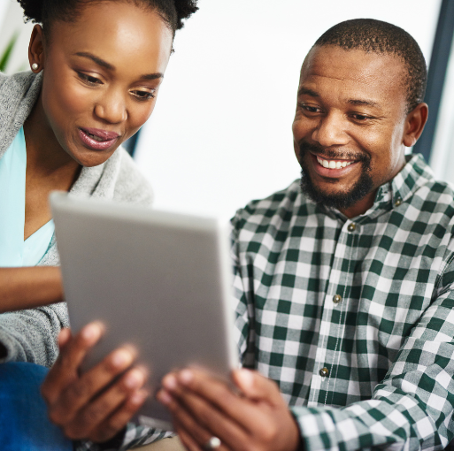 Two people smiling and looking at a tablet together, appearing engaged and focused. One is wearing a checked shirt and the other a light blue top, sitting close and sharing the device.