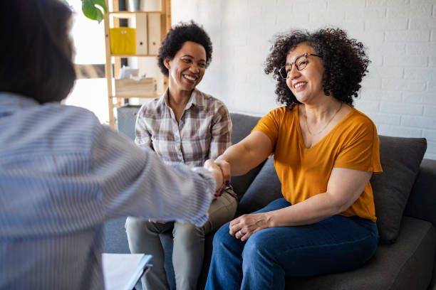 Two women sit on a couch, smiling as one shakes hands with a third person whose back is to the camera. The scene appears friendly and welcoming, suggesting a positive interaction in a behavioral health or counseling setting.