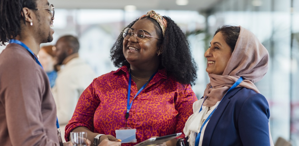 Three people wearing conference badges are smiling and conversing indoors, perhaps sharing insights about foster care. One person holds a glass and another holds a tablet as they engage in lively discussion. The setting is bright and modern, with blurred figures in the background.