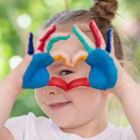 Young child with colored paint on hands forms a heart shape with fingers, smiling and looking through the heart. Lush greenery is blurred in the background.