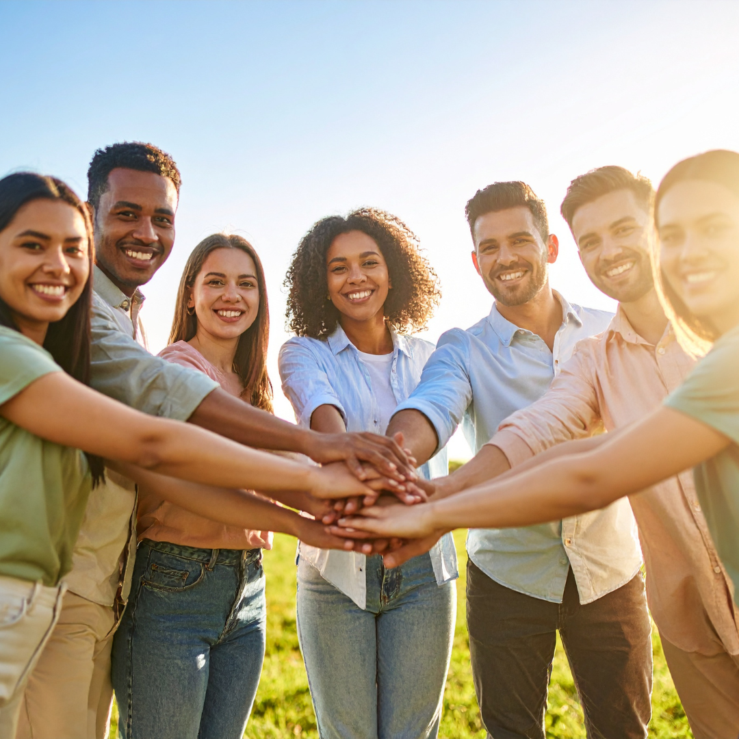 A group of smiling young adults stands in a circle outdoors, stacking their hands together in the center, showing teamwork and unity on a sunny day.