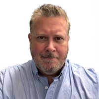 A middle-aged man with short, light hair and a beard, wearing a light blue collared shirt, is facing the camera against a plain white background.