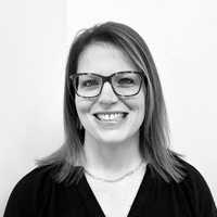 Smiling woman with straight shoulder-length hair, wearing glasses, a dark top, and a necklace, standing in front of a plain white wall. The photo is in black and white.