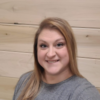 Smiling woman with long, straight, light brown hair wearing a grey top, standing in front of a light wooden plank wall.
